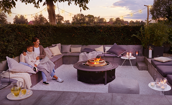 Gartensitzgruppe mit gepolsterten Bänken, zentraler Feuerstelle und Beleuchtung; eine Frau und ein Kind sitzen gemeinsam in Decken gehüllt