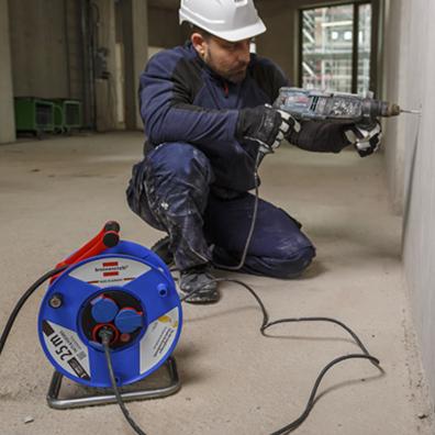 Ein Mann bohrt mit einem Bohrer in eine Wand und im Vordergrund sieht man eine Kabeltrommel von Brennenstuhl