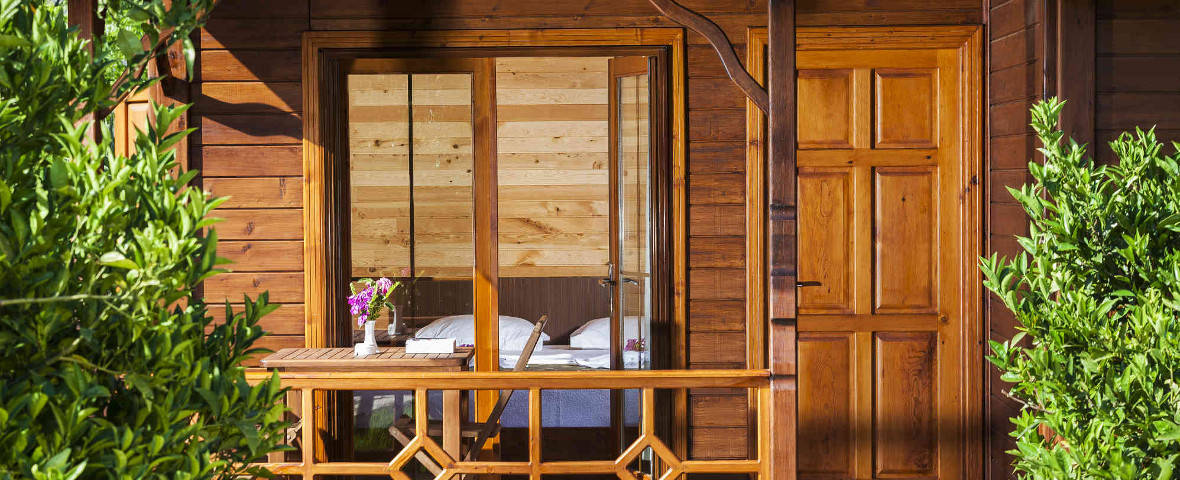Ein gemütliches Gartenhaus aus Holz mit überdachter Veranda und großen Fenstern gibt den Blick auf ein Schlafzimmer frei. Auf dem Tisch steht eine Blumenvase mit frischen Blumen