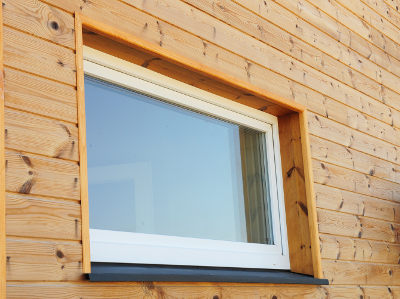 Nahaufnahme einer Hauswand aus Holz, in der der ein weißes Kunsstofffenster verbaut und mit einer schwarzen Fensterbank versehen ist
