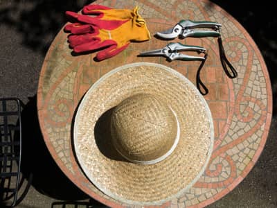 Ein runder Gartentisch mit Mosaikmuster, auf dem Tisch liegen ein Strohhut, zwei Gartenscheren und Gartenhandschuhe