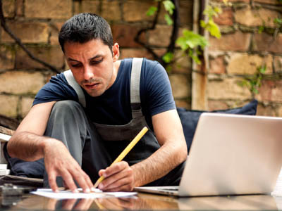 Mann in Arbeitskleidung schreibt mit Bleistift neben einem Laptop.