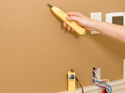 Foto einer Hand, die mit einem Ortungsgerät die Wand absucht, darunter ist eine offene Kabeldose mit herausgeführten Drähten und einem angeschlossenen Prüfgerät zu sehen