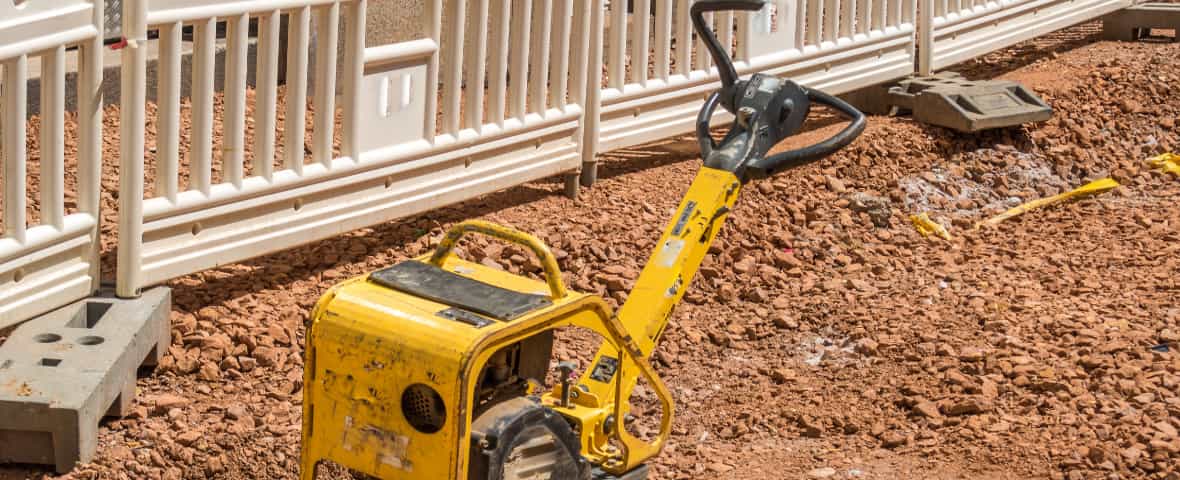 Gelbe Rüttelplatte auf einer Baustelle mit Schotter und Absperrung.