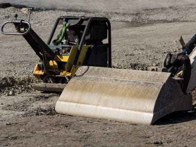 Baumaschinen auf Schotterfläche, Rüttelplatte und Baggerlöffel.