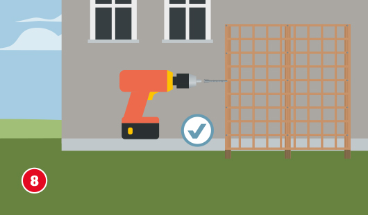 Ein Akkuschrauber wird verwendet, um eine fertige Rankhilfe aus Holzgittern an einer Hauswand zu befestigen. Die Rankhilfe steht stabil zwischen drei senkrechten Holzpfosten. Ein Häkchen-Symbol zeigt an, dass dieser Schritt erfolgreich abgeschlossen ist.