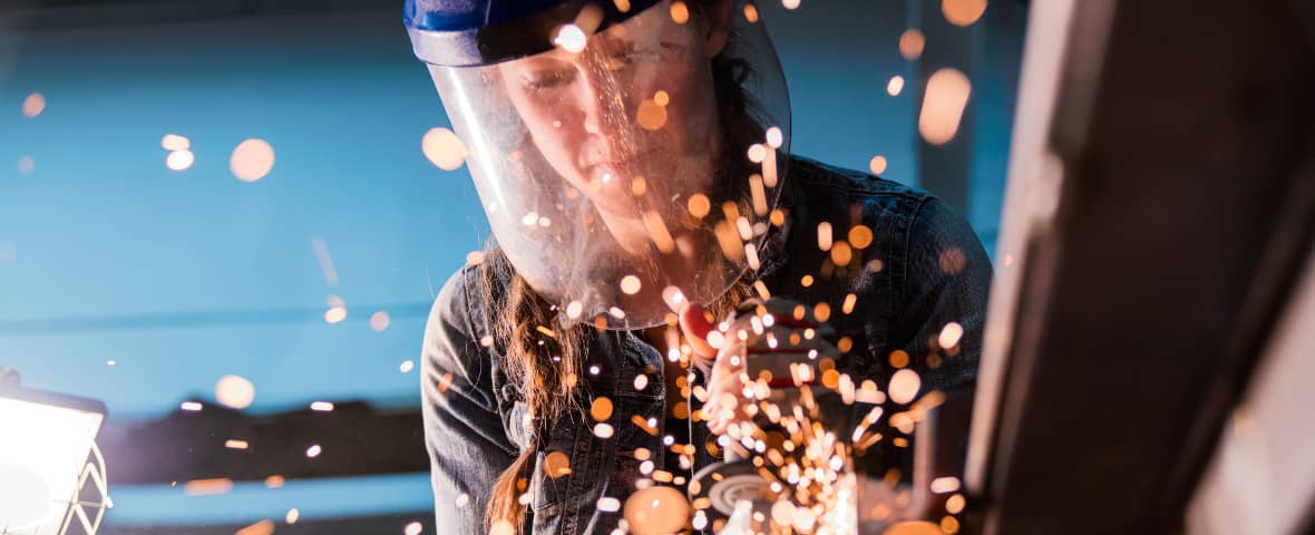 Eine Person trägt Schutzvisier und bearbeitet ein Metallstück mit einem Winkelschleifer. Funken fliegen in alle Richtungen.