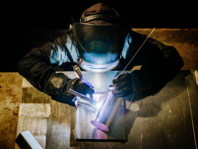 Eine Person mit Schweißhelm und Handschuhen führt eine präzise Schweißung an einem Metallrohr auf einem Arbeitstisch durch. Die Lichtbögen leuchten blau-violett, die Umgebung ist dunkel gehalten