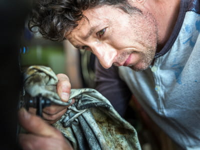 Ein Mann reinigt konzentriert ein Metallteil mit einem schmutzigen Lappen. Er trägt ein langärmliges Shirt und beugt sich über das Werkstück, das aus einem Schweiß- oder Werkstattkontext stammt
