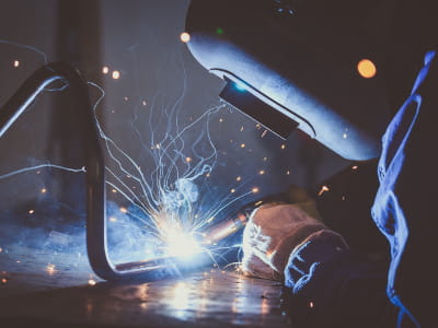 Eine Person mit Schweißhelm und Handschuhen führt eine Lichtbogenschweißung an einem gebogenen Metallrahmen aus. Blaues Licht, Funkenflug und Rauch begleiten die Arbeit im dunklen Werkstattumfeld