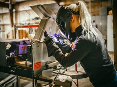 Eine Frau mit Schutzhandschuhen und Schweißhelm führt eine Schweißung an einem Werkstück in einer Werkhalle durch