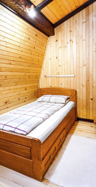 Ein kleines Schlafzimmer mit holzverkleideten Wänden und Dachschrägen ist mit einem Einzelbett aus Holz eingerichtet. Eine schlichte Lampe an der Decke sorgt für Beleuchtung
