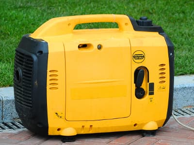 Ein kompakter gelber Inverter-Stromerzeuger mit Tragegriff steht auf Pflastersteinen neben einer Rasenfläche