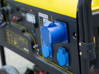 Eine Nahaufnahme zeigt die Steckdosenleiste eines mobilen Stromerzeugers mit gelbem Gehäuse. Zu sehen sind mehrere blaue Schutzkontakt- und CEE-Steckdosen sowie Bedienelemente und ein Rahmenbügel
