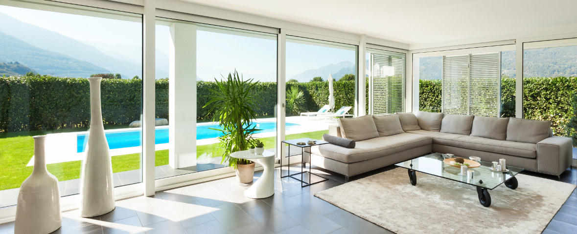 „Foto eines modernen Wohnzimmers mit bodentiefen Fenstern und Blick auf eine Terrasse mit Pool und Garten, der Raum ist mit einem großen, hellgrauen Ecksofa, einem Glastisch auf Rollen, hellen Deko-Vasen und einem Teppich ausgestattet