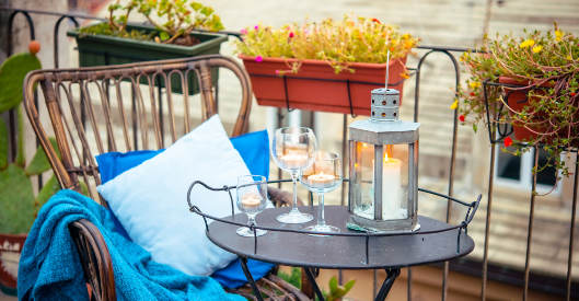 Foto von einem kleinen Metalltisch auf einem Balkon, auf dem Tisch stehen eine Lampe und Wein- und Wassergläser mit Kerzen, am Balkongeländer sind Blumenkästen befestigt und am Tisch steht ein Holzstuhl mit einer Decke und einem Kissen