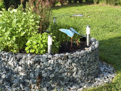 Ein bepflanztes Hochbeet mit Gabionen-Einfassung ist mit zwei kleinen Solarlampen ausgestattet. Zwischen den Kräutern steht eine dekorative blaue Metall-Schmetterlingsfigur