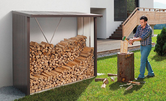 Ein Biohort Kaminholzlager an einer Hauswand, davor spaltet ein Mann Holz