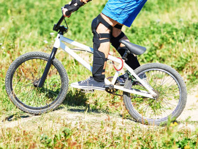 Ein Kind mit blauer kurzer Hose fährt ein weißes BMX-Fahrrad über einen Feldweg. Es trägt schwarze Schutzkleidung an Armen und Beinen