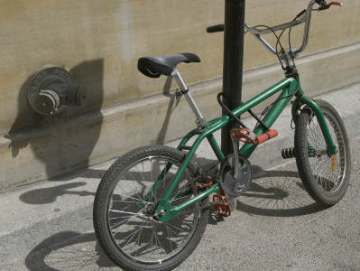 Ein grünes Kinderfahrrad ist an einem schwarzen Pfosten auf einem Gehweg angeschlossen. Es steht vor einer sandfarbenen Wand mit einem alten Wandhydranten auf der linken Seite