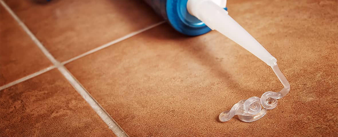 Nahaufnahme einer Kartuschenspritze, aus der transparente Silikonmasse in eine Fuge zwischen braunen Bodenfliesen aufgetragen wird