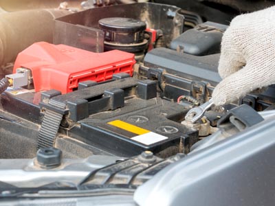 Nahaufnahme einer Autobatterie, an der von einer Hand in einem Arbeitshandschuh mit einem Schraubenschlüssein eine Schraube gelöst wird