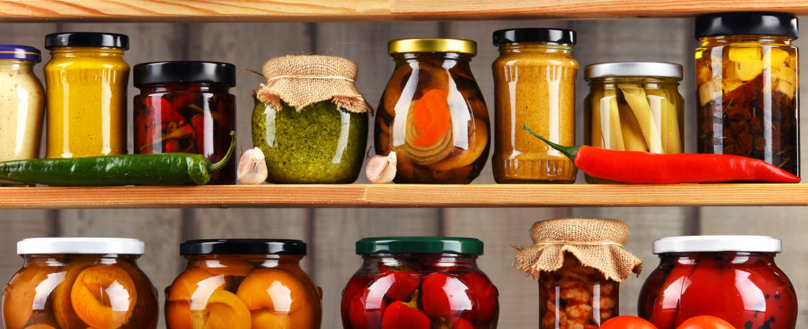 Auf einem Holzregal stehen zahlreiche Einmachgläser mit eingelegtem Obst, Gemüse und Saucen. Die Gläser sind bunt gefüllt und ordentlich nebeneinander aufgereiht.