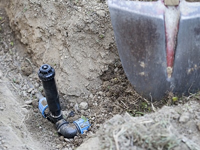 Nahaufnahme eines ausgehobenen Erdgrabens, in dem eine Sprinklerleitung verlegt wird, man sieht ein schwarzes Rohr mit blauen Verbindungsstücken und einen Sprinklerkopf, rechts im Bild lehnt eine Schaufel