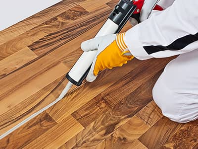 Foto wie eine Person in Arbeitskleidung und gelbem Handschuh, mit einer Kartuschenpistole weiße Dichtmasse in eine Fuge im Holzfußboden einbringt