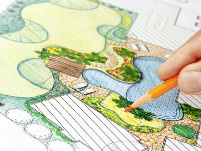 Eine farbig gestaltete Gartenplanungsskizze zeigt verschiedene Flächen, Wege und ein Wasserbecken. Eine Hand zeichnet Details mit einem orangen Buntstift ein