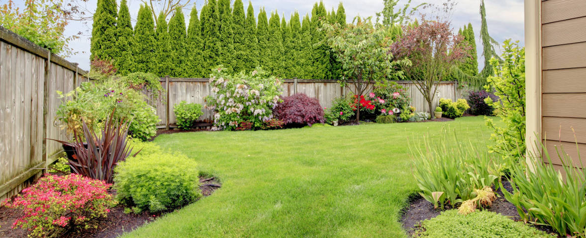 Ein gepflegter Garten mit grünem Rasen ist von bunten Ziersträuchern und Blumenbeeten eingerahmt. Im Hintergrund verläuft ein Holzzaun mit Sichtschutzhecke
