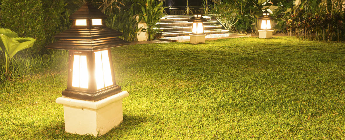 Eine Reihe stilvoller Gartenleuchten mit warmweißem Licht säumt einen gepflegten Rasenweg. Die Leuchten stehen auf Sockeln und beleuchten den Weg zu einer beleuchteten Treppe vor üppiger Bepflanzung