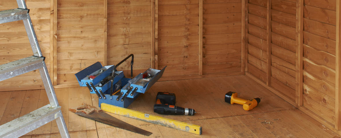 Innenansicht eines hölzernen Gartenhauses im Aufbau. Auf dem Boden liegen eine Werkzeugkiste, Akkuschrauber, Wasserwaage, Säge und Schrauben. Links steht eine Aluminiumleiter.