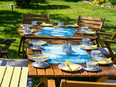 Zwei zusammengestellte Holztische im Garten, gedeckt im Porzellangeschirr. Um die Tische stehen mehrere Stühle aus Holz