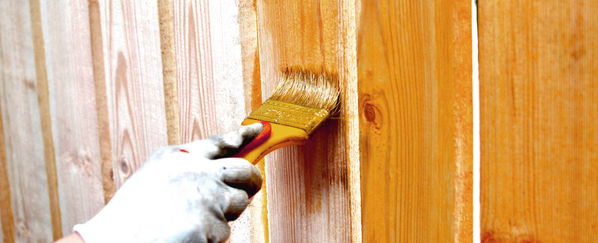 Eine Person trägt mit einem Pinsel Holzschutzmittel auf einen Gartenzaun auf, um das Holz vor Verwitterung und Verrottung zu schützen