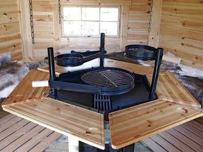 Der Innenraum einer Grillkota mit zentralem Grillplatz, umgeben von hölzernen Sitzbänken. An den Wänden befinden sich Fenster, die für natürliches Licht sorgen.