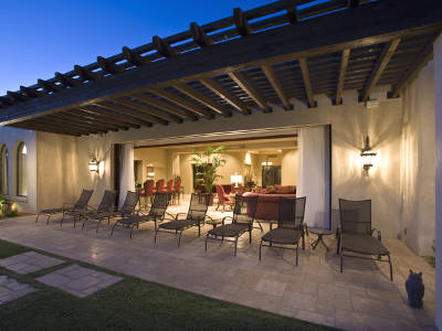 Eine überdachte Terrasse mit mehreren Liegestühlen ist dezent mit Wandleuchten beleuchtet. Der offene Wohnbereich dahinter ist hell erleuchtet und lädt zum Verweilen ein. Die Szene spielt in der Dämmerung