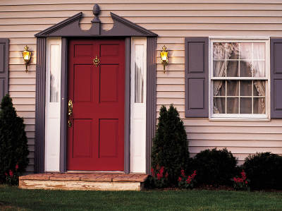 Außenansicht vom Eingangsbereich eines Hauses mit roter Haustür, die von grauen und weißen Zierleisten eingerahmt ist, links und rechts der Tür hängen Wandleuchten, daneben befinden sich Fenster mit Fensterläden und kleine Büsche im Vorgarten