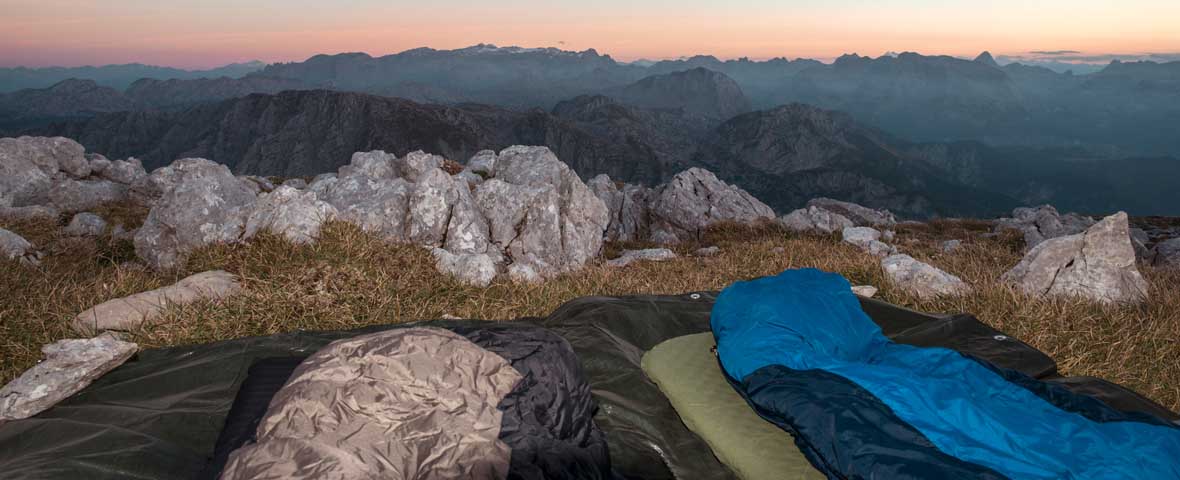 Foto von ausgebreiteten Schlafsäcken, die auf einer Wiese in felsigem Gelände in den Bergen liegen, im Hintergrund erstreckt sich eine Berglandschaft
