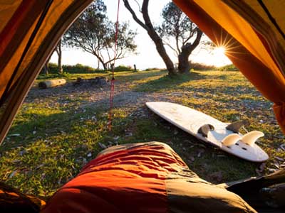 Blick aus dem Inneren eines Zeltes bei Sonnenaufgang, im Vordergrund liegt ein Schlafsack, draußen auf dem Gras steht ein Surfbrett