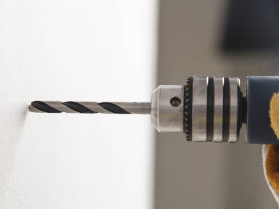 Eine Bohrmaschine mit eingespanntem Fliesenbohrer wird in Nahaufnahme an eine glatte Fliesenoberfläche angesetzt. Der Handwerker trägt einen orangefarbenen Arbeitshandschuh