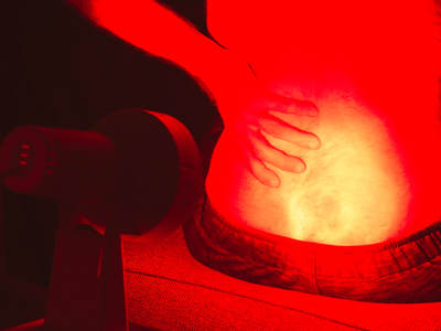 Eine Person bestrahlt ihren unteren Rücken mit einer eingeschalteten Rotlichtlampe. Die betroffene Stelle wird intensiv rot beleuchtet.