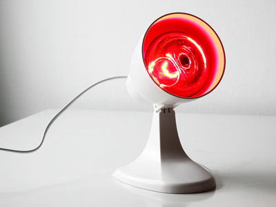 Eine eingeschaltete Rotlichtlampe steht auf einem weißen Tisch. Die Lampe strahlt intensiv rotes Licht ab und ist an eine Stromquelle angeschlossen.
