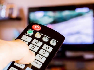 Eine Hand hält eine Fernbedienung, die auf einen eingeschalteten Fernseher gerichtet ist. Der Bildschirm zeigt ein unscharfes Bild.