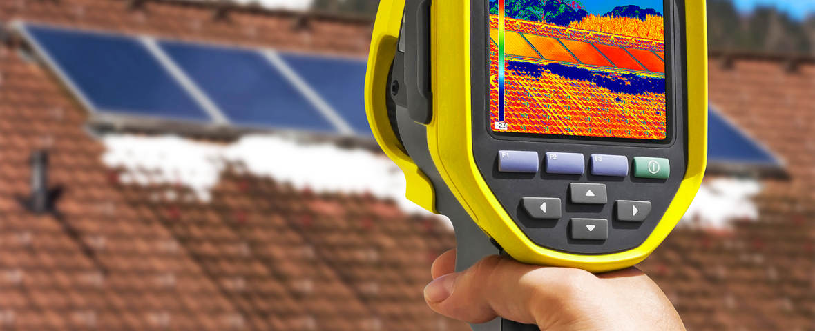 Eine Person hält eine Wärmebildkamera auf ein Dach mit Solarpaneelen gerichtet. Auf dem Display sind Temperaturunterschiede des Daches farbig dargestellt.