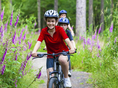 Drei Kindern fahren mit dem Fahrrad einen schmalen Waldweg entlang. Das vorderste Kind trägt einen Fahrradhelm und ein rotes Polo-Shirt. Umgeben sind sie von blühenden lila Blumen und dichtem Grün