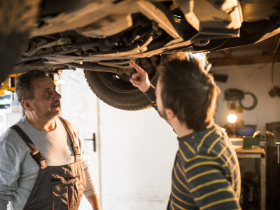 Zwei Männer stehen unter einem aufgebockten Auto in einer Werkstatt. Einer von ihnen zeigt auf einen Bereich im Unterboden, während der andere aufmerksam zuschaut
