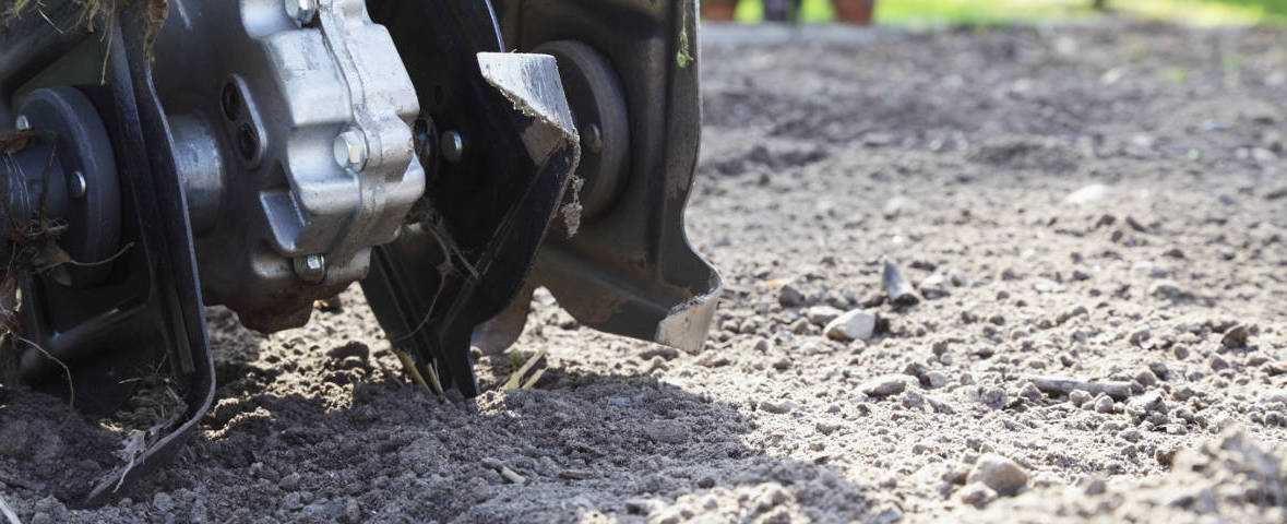 Die rotierenden Messer einer Motorhacke graben sich in trockene, bröckelige Erde. Die Aufnahme zeigt die Bodenbearbeitung aus der Nähe bei hellem Tageslicht