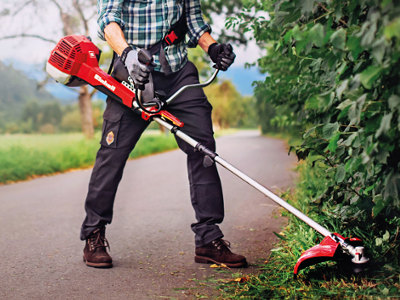 Eine Person mit Schutzkleidung und Tragegurt arbeitet mit einer roten Motorsense am Rand eines Weges. Die Motorsense wird entlang einer dichten Hecke geführt, um Rasenkanten und Wildwuchs zu trimmen
