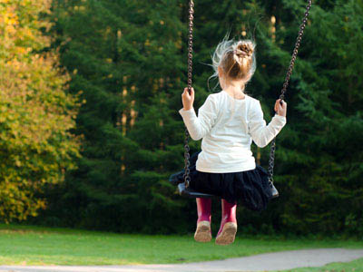 Ein kleines Mädchen schaukelt auf einer Schaukel, sie ist von hinten zu sehen, im Hintergrund stehen Bäume